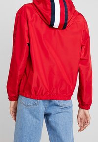 Giacca a vento rossa con cappuccio caratterizzato da strisce blu navy e bianche. Polsini ed orlo elasticizzati, texture liscia e vestibilità comoda.