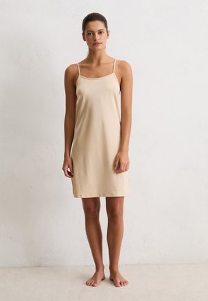 Vrouw die blootsvoets tegen een witte muur staat, gekleed in een beige slipdress zonder mouwen met dunne schouderbandjes, en recht vooruit kijkt met een neutrale uitdrukking.