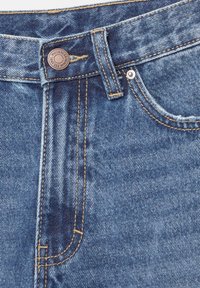 Gros plan sur un jean en denim bleu montrant la taille, la boucle de ceinture, un bouton en métal avec le texte de la marque et une poche avant avec rivet et couture jaune.