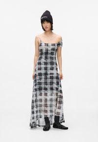 Kvinde i en sort-hvid ternet, one-shoulder maxi-kjole, sort hue, sorte sokker og sorte sko, der står op ad en hvid baggrund.