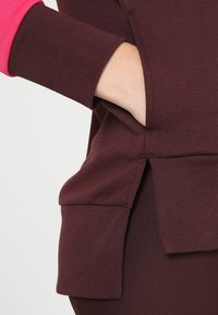 Sudadera burdeos con puños rosas, que presenta aberturas laterales y un tejido suave y texturizado. Vista en primer plano que enfatiza los detalles del bolsillo y las mangas.