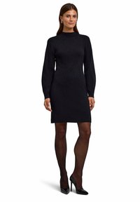Schwarzes Strickkleid mit langen Ärmeln, hohem Kragen und taillierter Silhouette. Kombiniert mit schwarzen High Heels und transparenten Strumpfhosen.