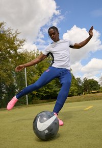 Nike focista fehér és fekete rövid ujjú pólóban, kék nadrágban, logóval, rózsaszín lábbeliben, egy fekete Nike labdát rúg a zöld fűre.