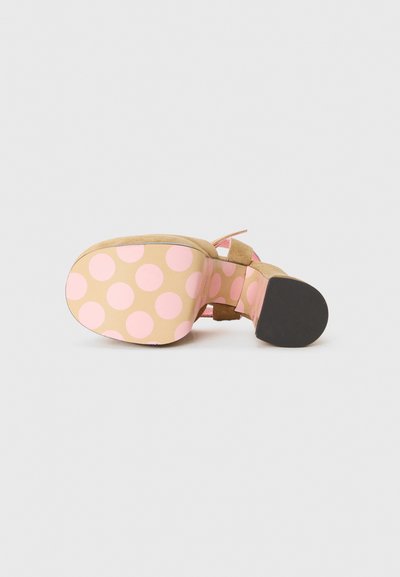 Beige mockasklackar med ett rosa prickigt mönster på sulan. Har en blockklack och en vristrem med en rosettdetalj.