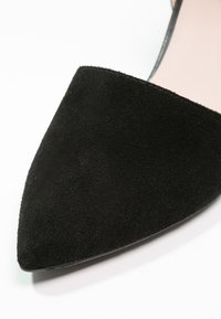 Chaussure plate en daim noir à bout pointu avec une fine anse autour de la cheville et une semelle intérieure rose clair, présentée sur un fond blanc.