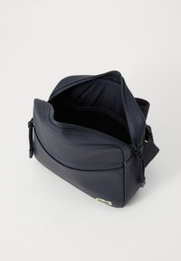 Geantă de culoare albastru marin, cu textură moale, cu închidere cu fermoar, căptușeală interioară din plasă și buzunar frontal. Prezintă un logo Lacoste pe lateral.