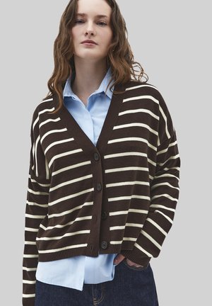 Donna che indossa un cardigan marrone e bianco a righe con bottoni sopra una camicia azzurra e jeans blu scuro, in piedi davanti a uno sfondo grigio.