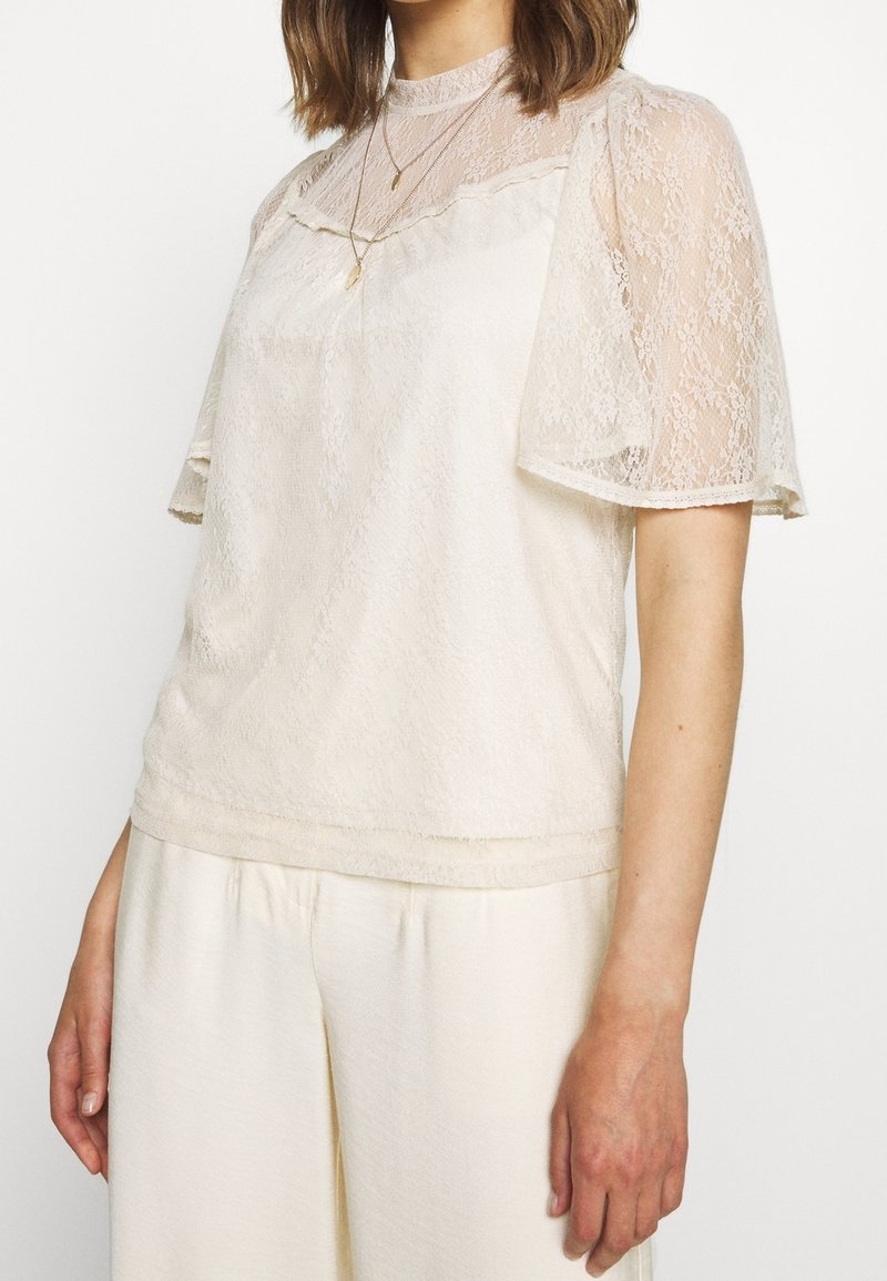 Femme portant un chemisier en dentelle crème avec des manches courtes transparentes et un pantalon large crème, accessoirisée d'un délicat collier en or.