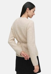 Sweter dziergany w miękkim beżowym kolorze, o dopasowanym kroju, z długimi rękawami i paskiem w talii. Posiada prążkowane mankiety oraz fakturowany materiał.