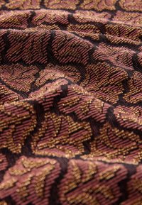 Textil s černým podkladem, který má strukturovaný tkaný vzor s jemně růžovými a zlatými akcenty, znázorňující motiv listu.