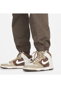 Hnědé a béžové vysoké tenisky s bílým logem Nike, obuté s hnědými joggingovými kalhotami na bílém pozadí.