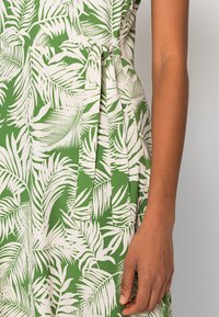 Arm of person wearing green and white tropical leaf-patterned dress with side tie detail against a plain background.