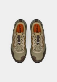 Μπεζ αθλητικά παπούτσια με καφέ λεπτομέρειες, υφαντό επάνω μέρος, λαβή από καουτσούκ και ορατό λογότυπο. Διαθέτουν απορρόφηση κραδασμών και κλείσιμο με κορδόνια.