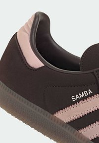 Zapatilla de ante negro con detalles en rosa, que presenta tres franjas horizontales, una suela de goma texturizada y el logo "SAMBA" en el lateral.
