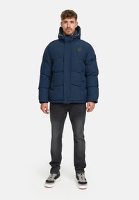 Mężczyzna stoi w granatowej kurtce puchowej z kapturem, czarnych dżinsach i czarno-białych sneakersach na tle białego tła.