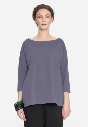 Kobieta o jasnej karnacji i brązowych włosach, ubrana w luźną fioletową bluzkę z długim rękawem i czarne spodnie, nosząca na lewej ręce zielone, brązowe i fioletowe bransoletki.