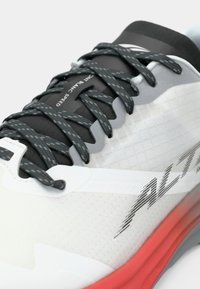 Chaussure de sport avec une tige perforée blanche, des accents gris, des lacets noirs et une semelle rouge. Présente un logo angulaire et un design épuré.