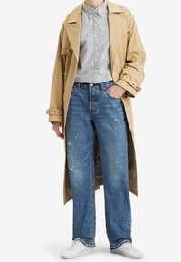 Gabardina beis sobre una camisa de botones a rayas, combinada con jeans de pierna ancha de color azul claro con detalles desgastados y zapatillas blancas.