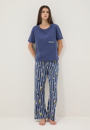 Vrouw staat op blote voeten, draagt een blauw T-shirt met de tekst "PEANUTS" en blauw gestreepte pyjamabroek met Peanuts-figuren.