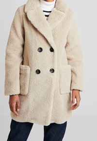 Abrigo beige de doble botonadura con una tela texturizada y esponjosa, gran solapa con muesca y dos bolsillos parche frontales. Botones negros que acentúan el diseño.