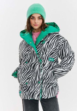 Młoda kobieta w czarno-białej kurtce puchowej w zebrze z zielonym wykończeniem, w zielonej czapce, różowym swetrze i ciemnych dżinsach, pozująca pewnie.