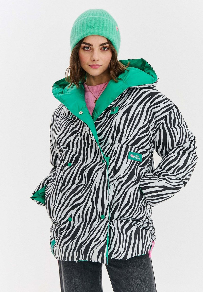 Mladá žena v čierno-bielom zebrovom pufrovom kabáte so zeleným podšíváním, so zelenou čiapkou, ružovým sveterom a tmavými džínami, chtiacou se fotiť s dôverou.