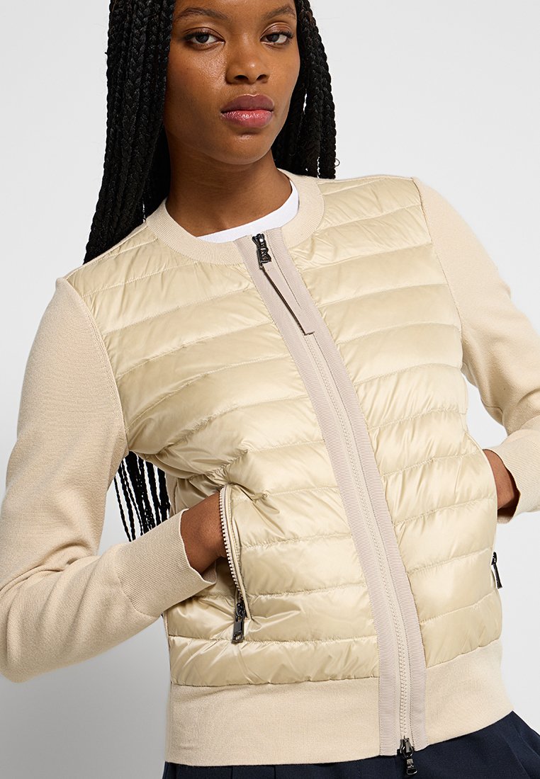 Frau mit geflochtenem Haar, die eine beige gesteppte Zip-Jacke trägt, mit den Händen in den seitlichen Reißverschlusstaschen, darunter ein weißes Hemd und eine dunkelblaue Hose.