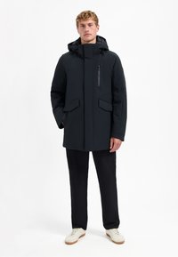 Zwarte waterdichte jas met capuchon, ritszak op de borst en voorzakken met klep. Heeft een rechte snit en een zachte textuur.