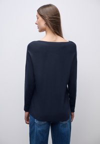 Marineblaues Langarmoberteil aus weichem Stoff, mit weitem Ausschnitt und lässiger Passform, kombiniert mit blauen Jeans. Glatte Textur, minimales Design.