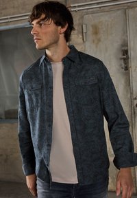 Granatowa koszula flanelowa z geometrycznym czarnym wzorem, zapięta na guziki, noszona na jasnoszarym T-shircie, w połączeniu z niebieskimi dżinsami.