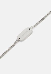 Pulsera de cadena de plata que presenta una etiqueta alargada con marca grabada, superficie lisa y un cierre sencillo.