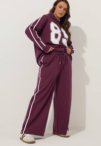 Burgunderfarbener Hoodie mit übergroßer weißer Nummer 85, kombiniert mit passenden weiten Sweatpants mit weißen Seitenstreifen. Weicher Stoff, lässige Passform.