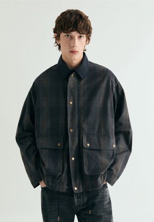 Chaqueta de cuadros negros con botones de presión dorados, bolsillos en el pecho y cuello con solapa. La tela tiene una textura suave con variados tonos oscuros.