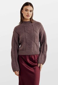 Rövidített, cable-knit pulóver mályva színben, magas nyakkal és laza ujjakkal. Bordó selyem szoknyára van húzva. Texturált, meleg anyagból készült.