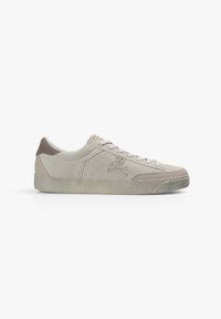 Zapatillas bajas beige con diseño de cordones, acentos de ante, detalles de perforación y un logo de calavera y huesos cruzados en el lateral.