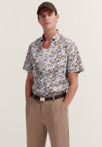 Kurzärmliges Hemd mit Knopfleiste, das ein blaues, braunes und cremefarbenes Blumendesign aufweist. Kombiniert mit beigen Hosen und einem braunen Gürtel.