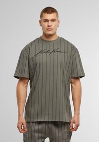 Camiseta de manga corta, en verde oliva, con rayas verticales negras y un gráfico distintivo en la parte frontal. Confeccionada en tela suave, de corte relajado.