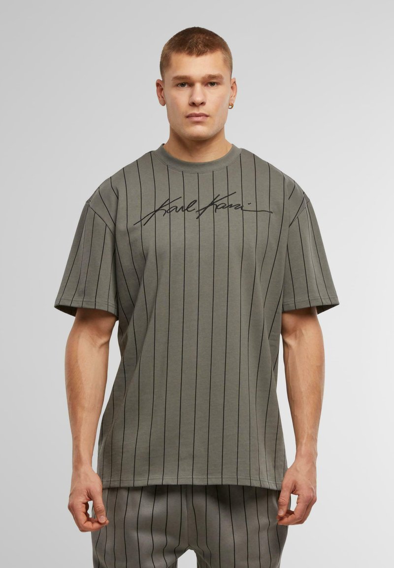 Camiseta de manga corta, en verde oliva, con rayas verticales negras y un gráfico distintivo en la parte frontal. Confeccionada en tela suave, de corte relajado.