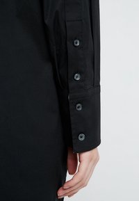 Detailní záběr ruky vedle černého rukávu košile se čtyřmi černými knoflíky na manžetě na světlém pozadí.