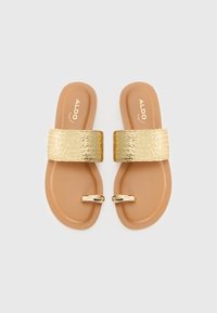 Goldene, gewebte Riemchensandalen mit einer flachen, tanfarbenen Sohle und dekorativen goldenen Beschlägen. Der Riemen weist ein strukturiertes Muster auf.