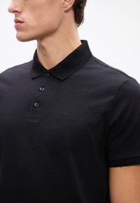 Mladý muž nosí čiernu polokošeľu s krátkym rukávom so štyrmi čiernymi gombíkmi a malým vyrazeným logom "cK" na hrudi.