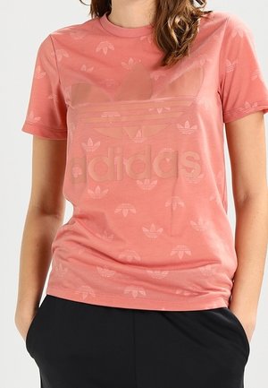 Femme portant un t-shirt rose Adidas avec un grand logo et de petits motifs trèfle, les mains dans les poches d’un pantalon noir.