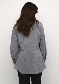Persona con lunghi capelli castani, che indossa una camicia button-up a righe sottili bianco e nera aderente e pantaloni neri, rivolta verso uno sfondo chiaro e uniforme.