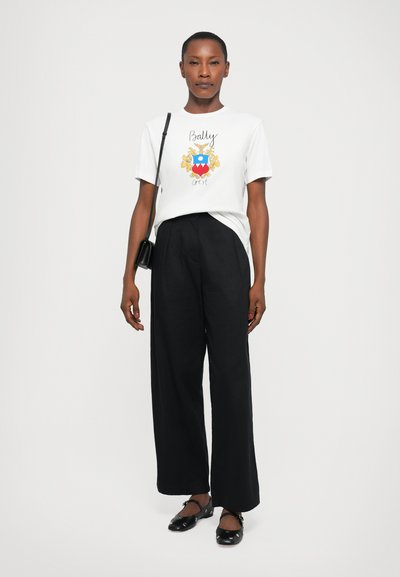 White cotton t-shirt with colorful Bally crest print, paired with loose black trousers and black shoes with decorative hardware accents.