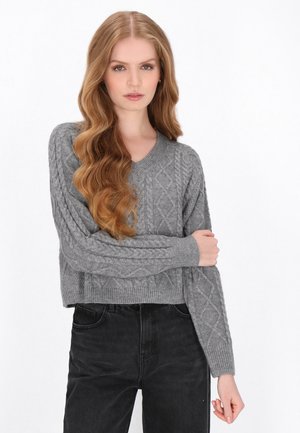 Ung kvinde med langt bølget hår iført en grå kabelstrikket sweater og sorte bukser, står med en arm krydset over brystet.