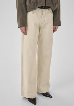 Persona che indossa pantaloni beige a gamba larga, scarpe nere a punta, maglia marrone a maniche lunghe, in piedi davanti a uno sfondo chiaro e uniforme.