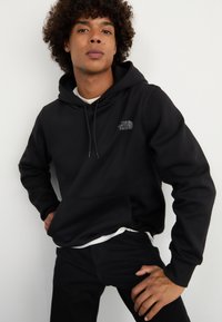 Felpa nera con cappuccio e tasca frontale, dotata di un logo grigio sul lato sinistro del petto. Tessuto morbido, vestibilità comoda, abbinata a jeans neri.