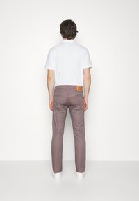 Man som bär en vit polo, mauve smal passform jeans med bakfickor och vita sneakers, står vänd mot en enkel vit vägg.