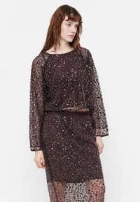 Bruine jurk met kralen en doorschijnende lange mouwen, met een aansluitend lijfje en een losse taille, versierd met verschillende sprankelende details.