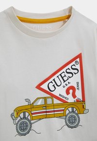 Weißes Baumwoll-T-Shirt mit einem gelb abgesteppten Kragen. Mit einer Grafik eines gelben Pickup-Trucks und einem roten Guess-Logo in Diamantform.